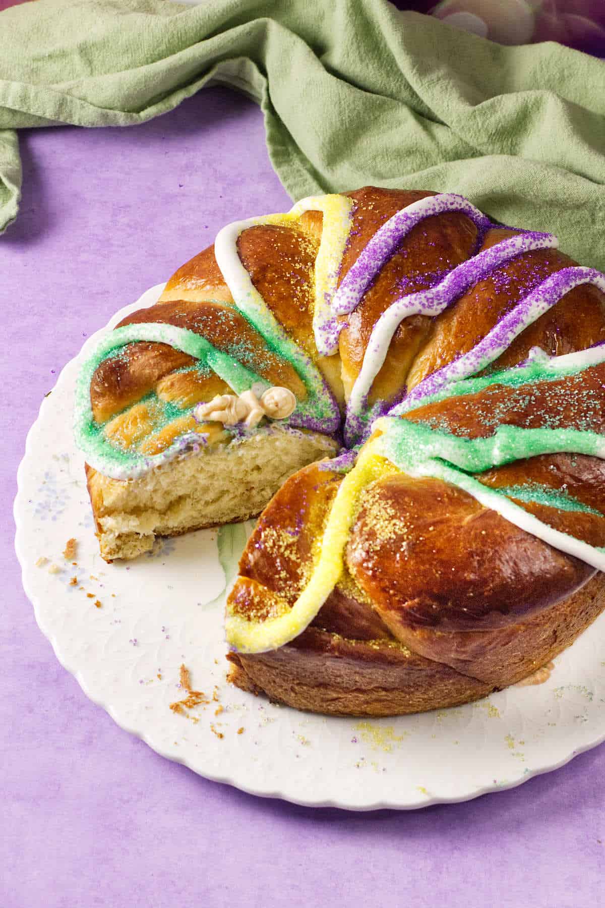 A round Mardi Gras King Cake with colorful sugar and icing in purple, yellow, and green sits on a white plate. One slice is cut out, revealing a small plastic baby inside. A green cloth rests in the background on a purple surface.