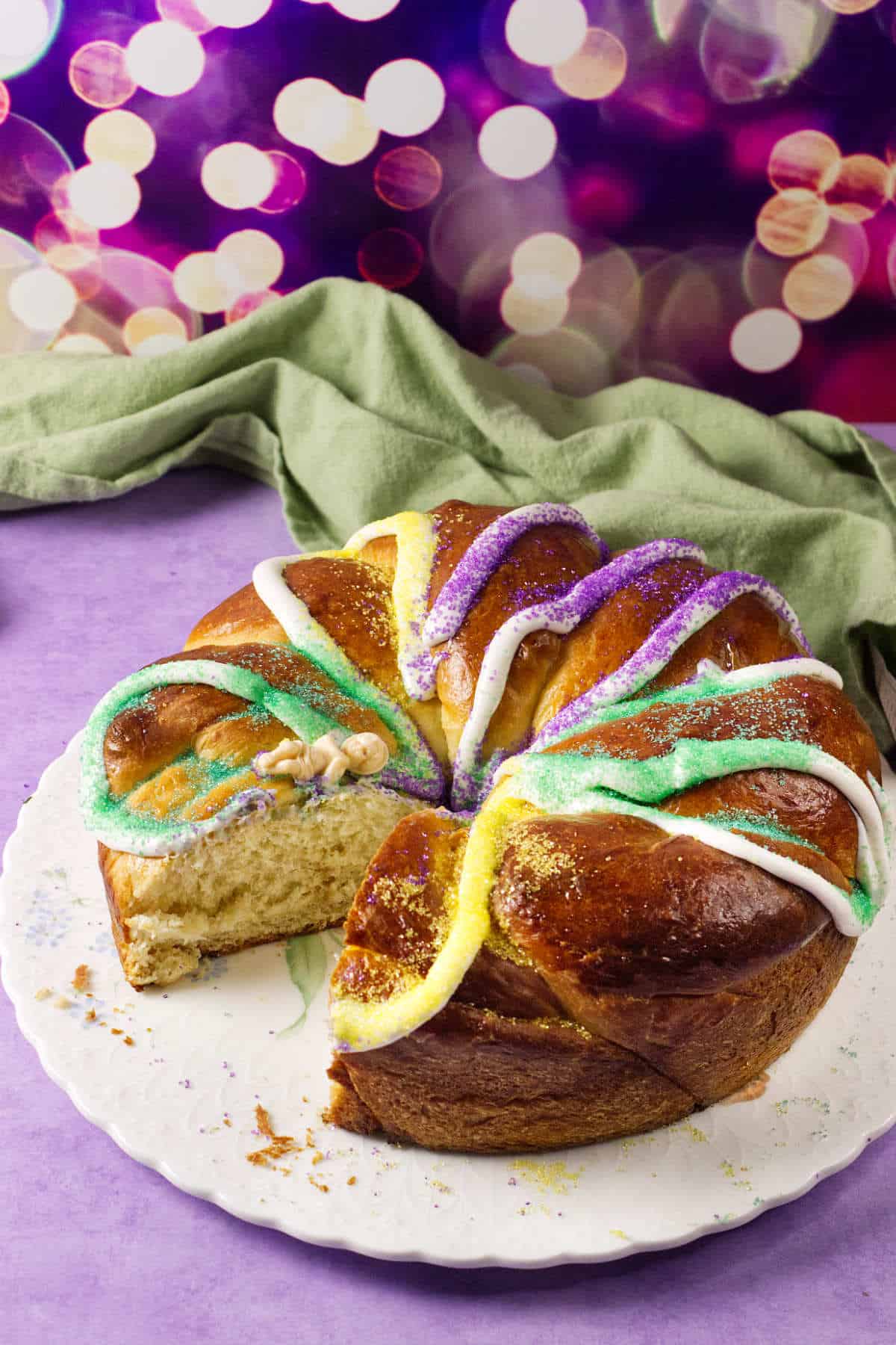 A round Mardi Gras king cake with a slice missing, topped with white icing and green, yellow, and purple sugar, sits on a white plate with a green cloth in the background and festive purple lights behind.