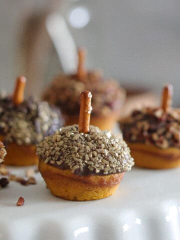 Mini acorn-shaped treats made from mini pumpkin muffins with chocolate-dipped tops, coated in chopped nuts or sprinkles, and pretzel stick stems, arranged on a white cake stand.