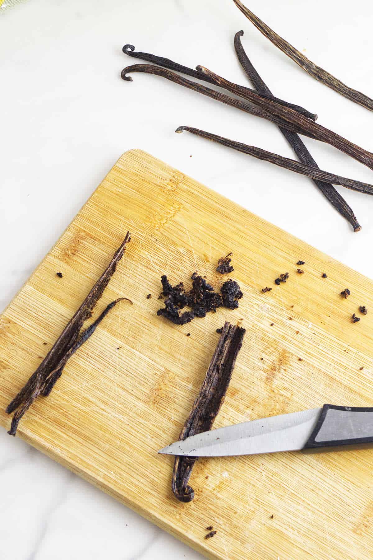 A wooden cutting board with split vanilla beans and scraped seeds, perfect for baking vanilla bean scones. A small pile of vanilla seeds is in the center, with a knife nearby and whole vanilla pods on a white surface.