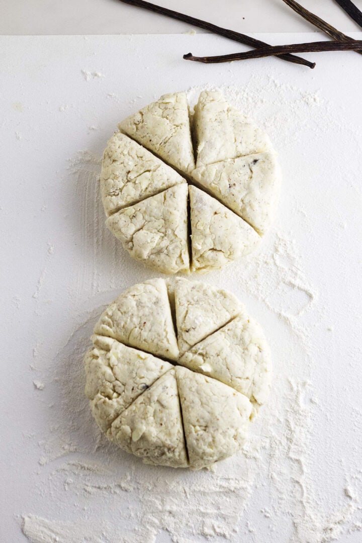 Two rounds of unbaked vanilla bean scone dough, each cut into six wedges, rest on a floured surface. Whole vanilla beans are visible near the top right corner.