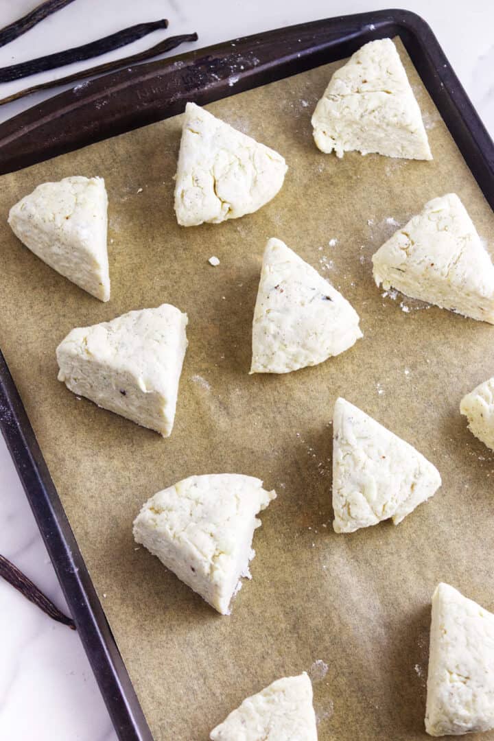 Unbaked, triangular vanilla bean scones are arranged on a parchment-lined baking sheet, ready to be baked. Vanilla beans rest in the top left corner.