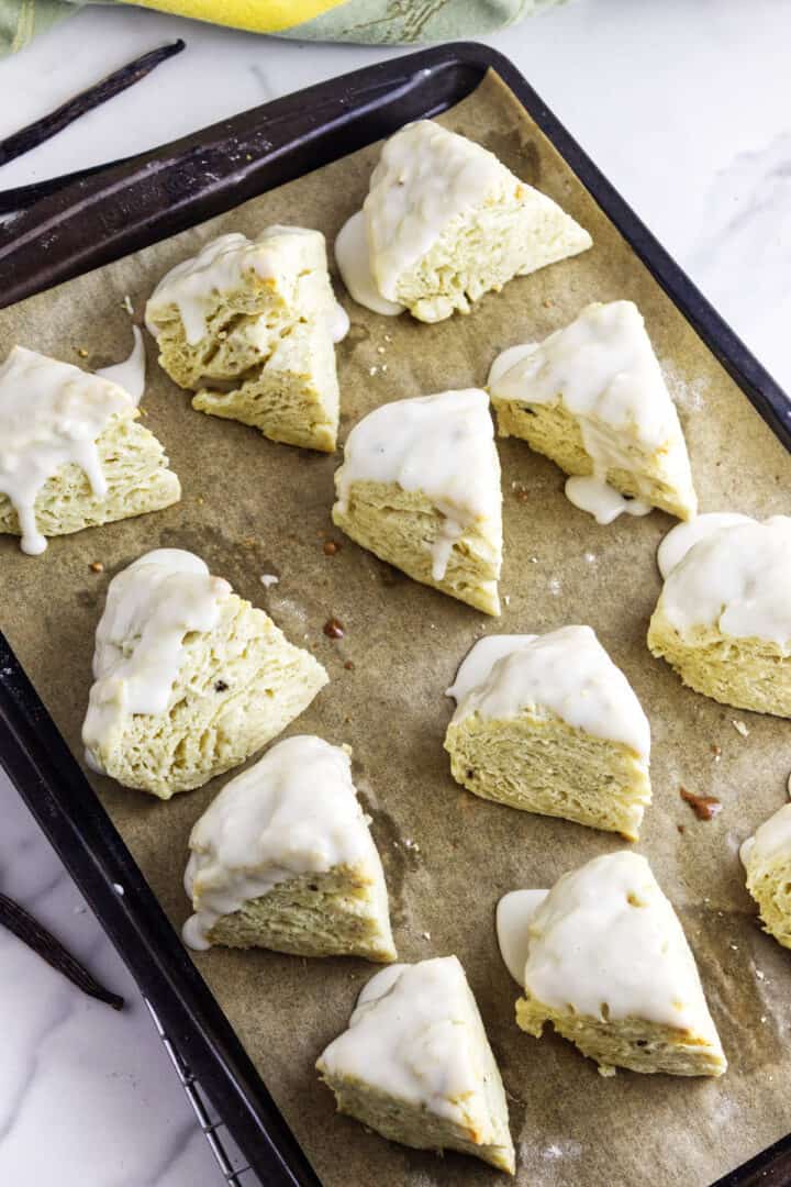 Freshly baked vanilla bean scones with a light glaze are arranged on a parchment-lined baking sheet, ready to serve. Some glaze drips down the sides, and the scones look fluffy and golden, set on a marble countertop.
