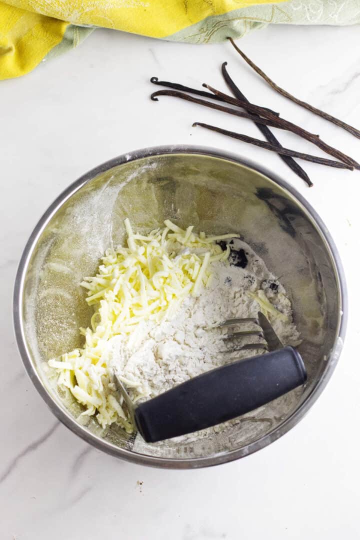 A metal bowl with flour and grated butter, a pastry cutter inside, sits on a white surface—ingredients coming together for vanilla bean scones. Nearby are whole vanilla beans and a yellow kitchen towel.