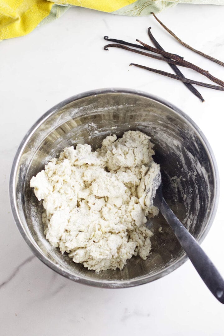 A metal mixing bowl filled with partially mixed vanilla bean scone dough and a black spatula sits on a white surface. Three vanilla bean pods and a yellow cloth are in the background.