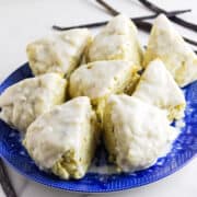 A blue plate holds eight triangular vanilla bean scones topped with a shiny white glaze. Vanilla beans are placed beside the plate on a white surface.