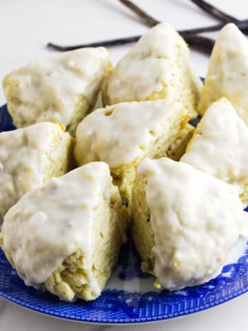 A blue plate holds eight triangular vanilla bean scones topped with a shiny white glaze. Vanilla beans are placed beside the plate on a white surface.