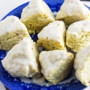 A blue plate holds several triangular vanilla bean scones topped with white icing, arranged in a circle on a light surface. Vanilla beans are visible in the background.