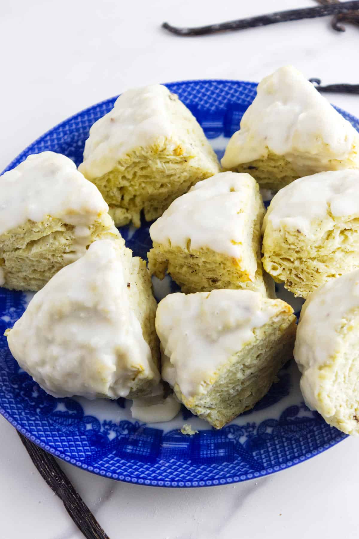 A blue plate holds several triangular vanilla bean scones topped with white icing, arranged in a circle on a light surface. Vanilla beans are visible in the background.