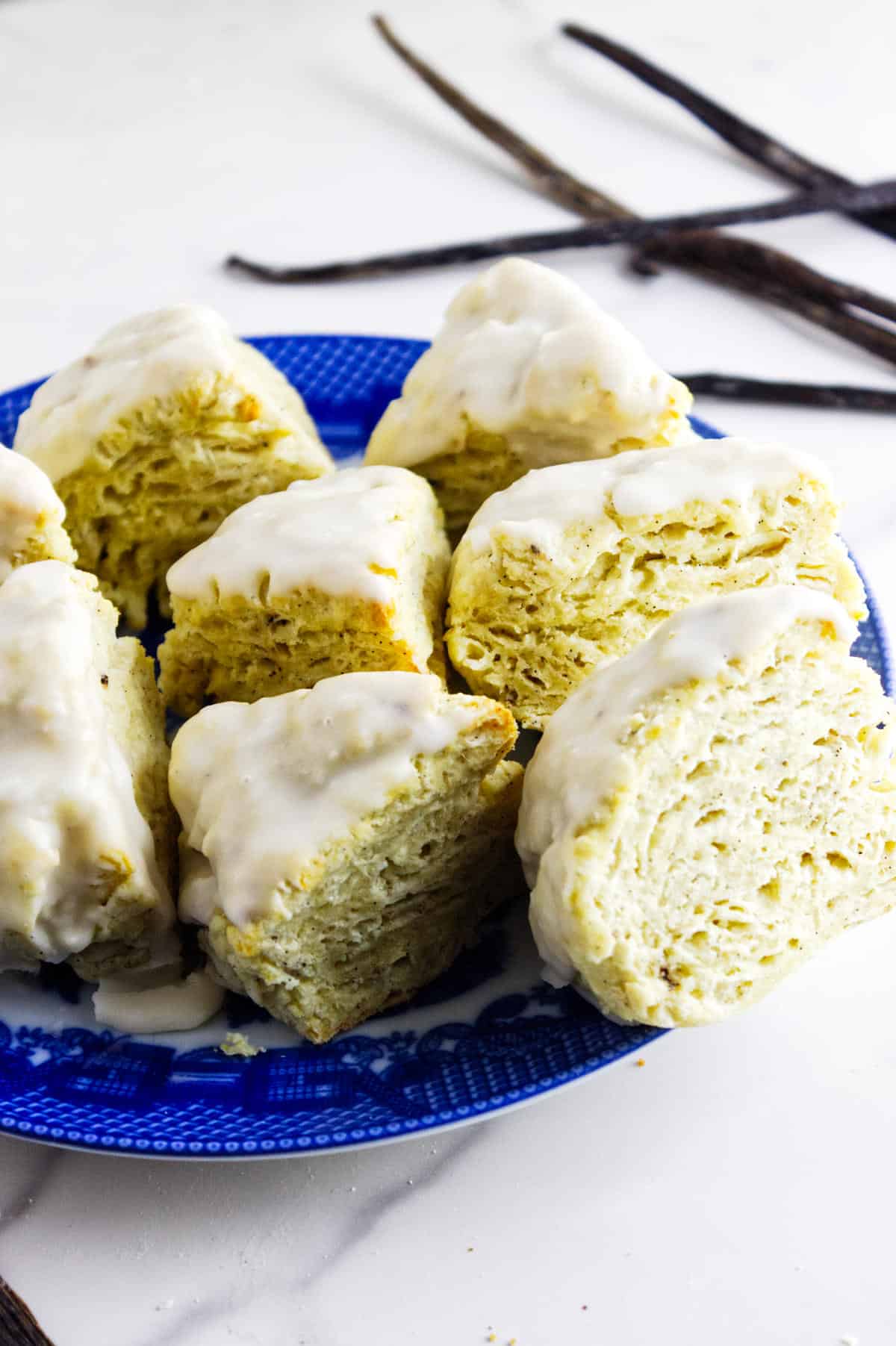 A blue plate holds several vanilla bean scones with a smooth vanilla glaze, stacked and arranged on a white surface. In the background, some vanilla bean pods are visible.