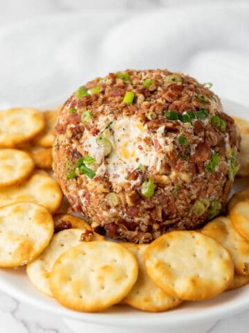 A pecan bacon cheeseball coated with chopped nuts and green onions sits on a white plate, surrounded by a ring of round, golden crackers. The background is softly blurred with a white cloth.