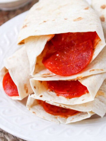 Three flour tortillas filled with slices of pepperoni, stacked on a white plate and ready to eat—these tasty pepperoni pizza roll ups bring classic flavors to every bite.