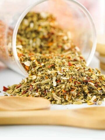 A glass jar tipped over, spilling a colorful pizza seasoning blend of dried herbs and spices onto a white surface, with wooden spoons nearby in the foreground.
