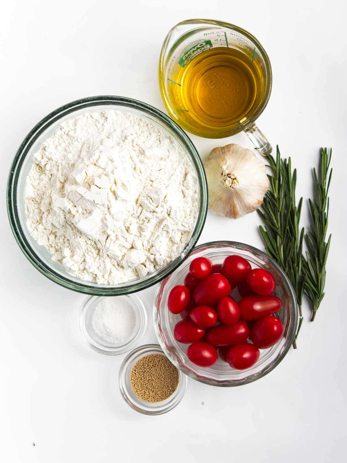 A flat lay of ingredients for roasted garlic focaccia on a white surface: a bowl of flour, cherry tomatoes, olive oil, a head of garlic, rosemary sprigs, and small bowls of salt and dry yeast.