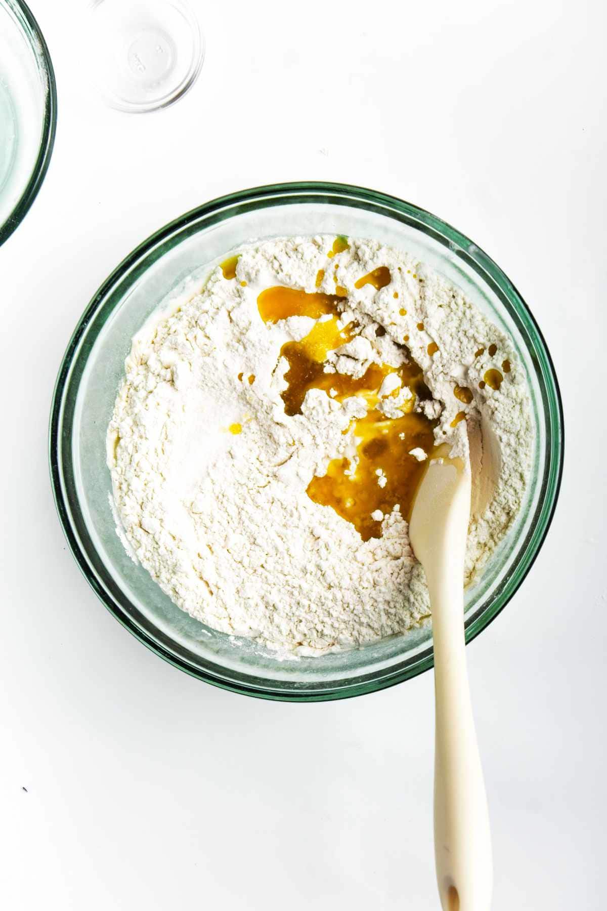 A glass bowl containing flour with a pool of vanilla extract and a spatula resting inside, set on a white surface-perfect for prepping dough for roasted garlic focaccia.
