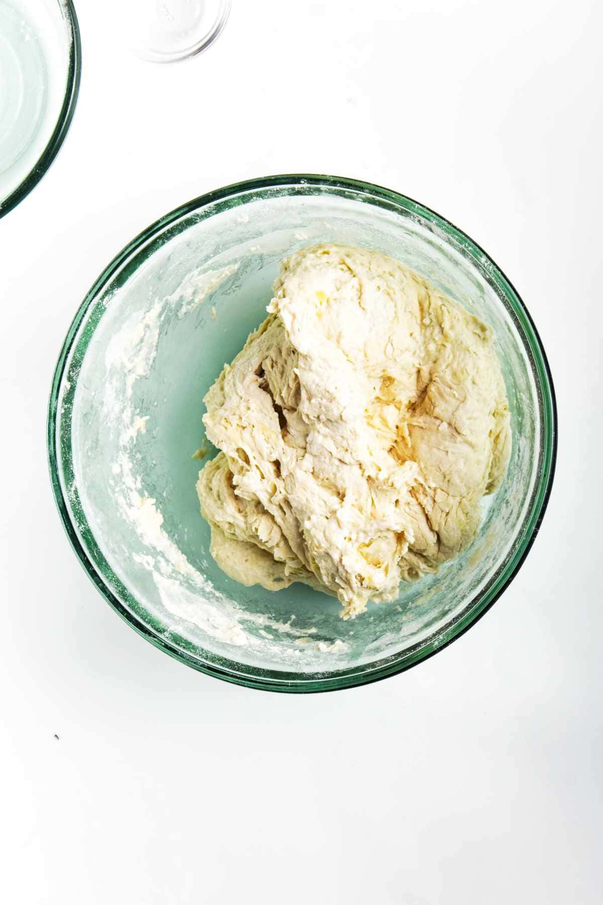 A glass bowl containing a ball of raw, partially mixed roasted garlic focaccia dough sits on a white surface. Some flour is visible on the sides of the bowl.
