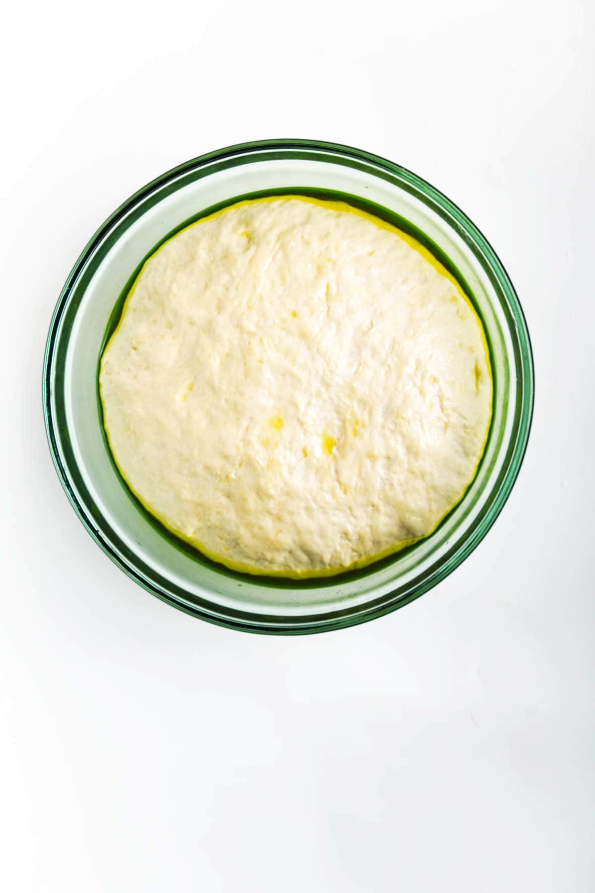 A glass bowl filled with risen roasted garlic focaccia dough sits on a white surface. The dough is smooth, puffy, and lightly coated with oil around the edges.