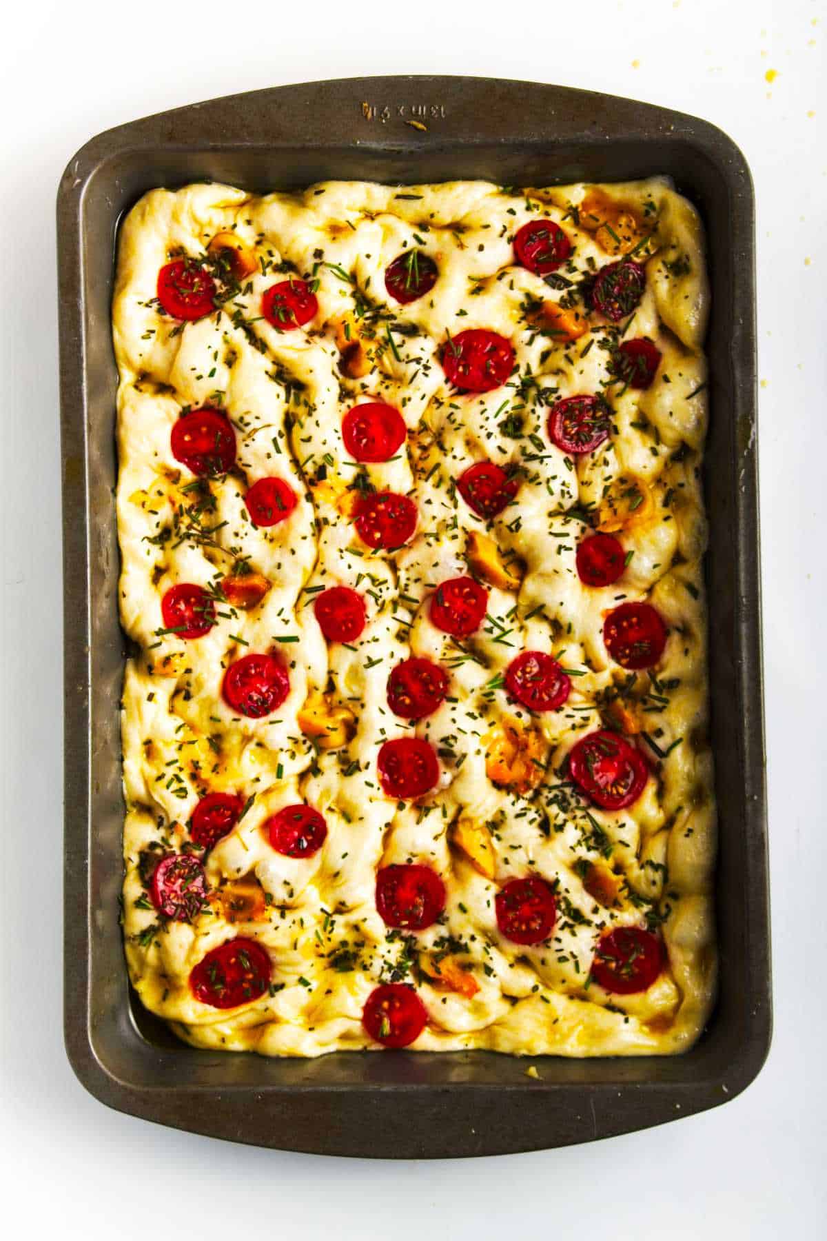 A rectangular baking pan filled with golden-brown roasted garlic focaccia, topped with cherry tomato slices, herbs, and tender roasted garlic cloves.