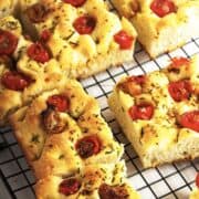 Golden slices of roasted garlic focaccia topped with cherry tomatoes and herbs rest on a black wire cooling rack. The bread looks fluffy inside with a crispy, browned crust.