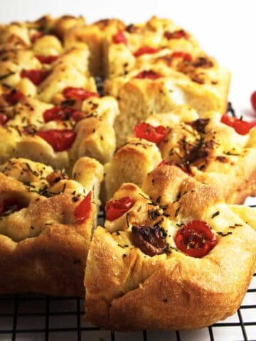 A golden-brown roasted garlic focaccia topped with cherry tomatoes and herbs, cut into squares and cooling on a black wire rack. A sprig of rosemary and a cherry tomato rest on the white surface beside it.