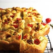 A sliced loaf of golden-brown roasted garlic focaccia bread topped with cherry tomatoes and herbs sits on a black cooling rack, with sprigs of rosemary and tomatoes nearby on a white surface.