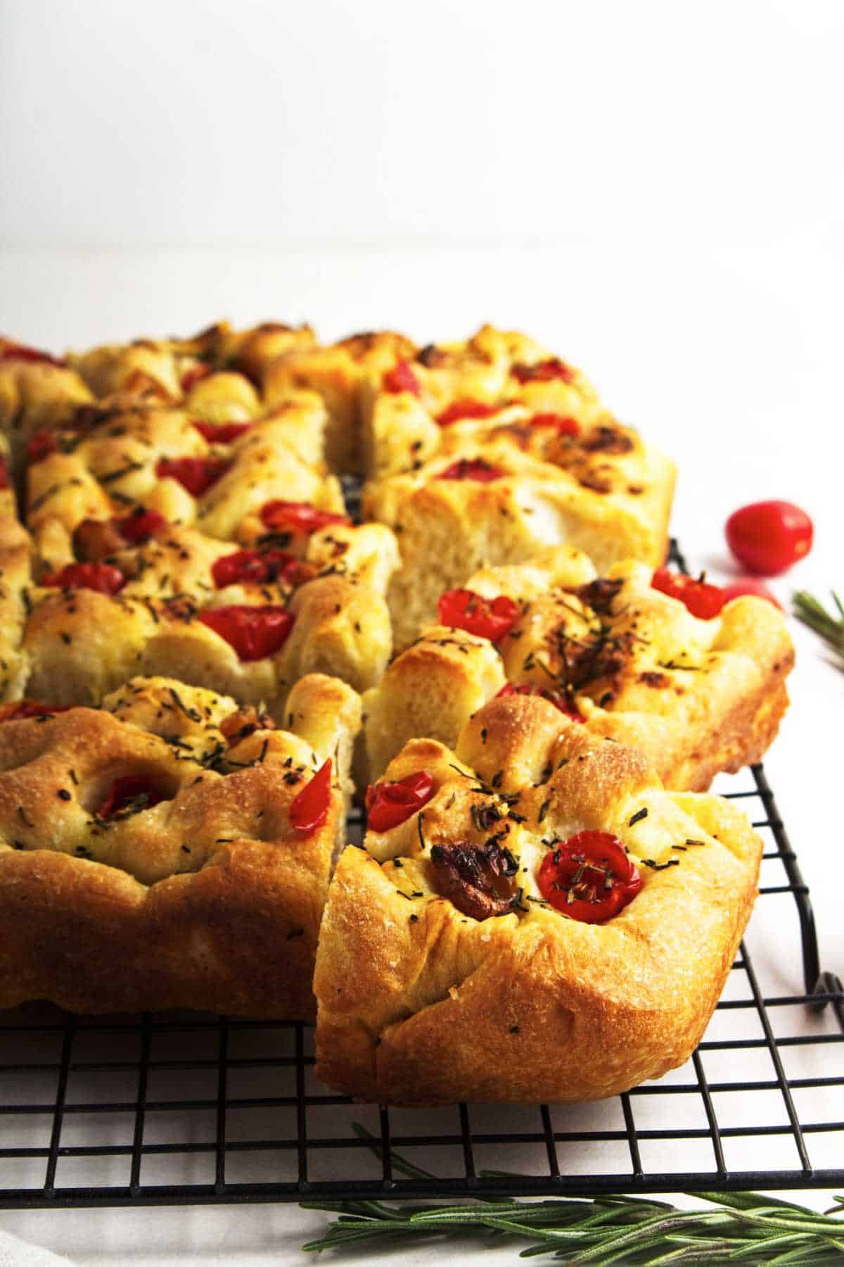 A sliced loaf of golden-brown roasted garlic focaccia bread topped with cherry tomatoes and herbs sits on a black cooling rack, with sprigs of rosemary and tomatoes nearby on a white surface.