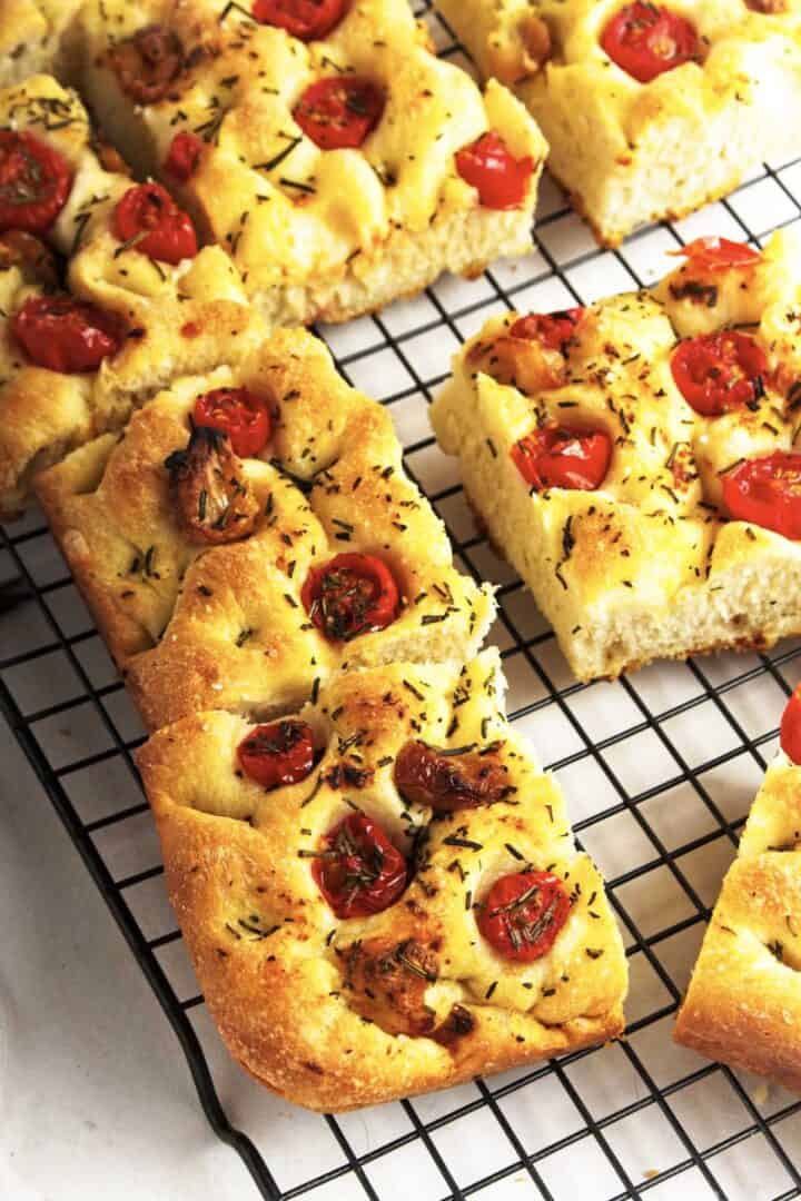 Freshly baked roasted garlic focaccia topped with cherry tomatoes and herbs, cut into rectangular pieces and cooling on a wire rack.