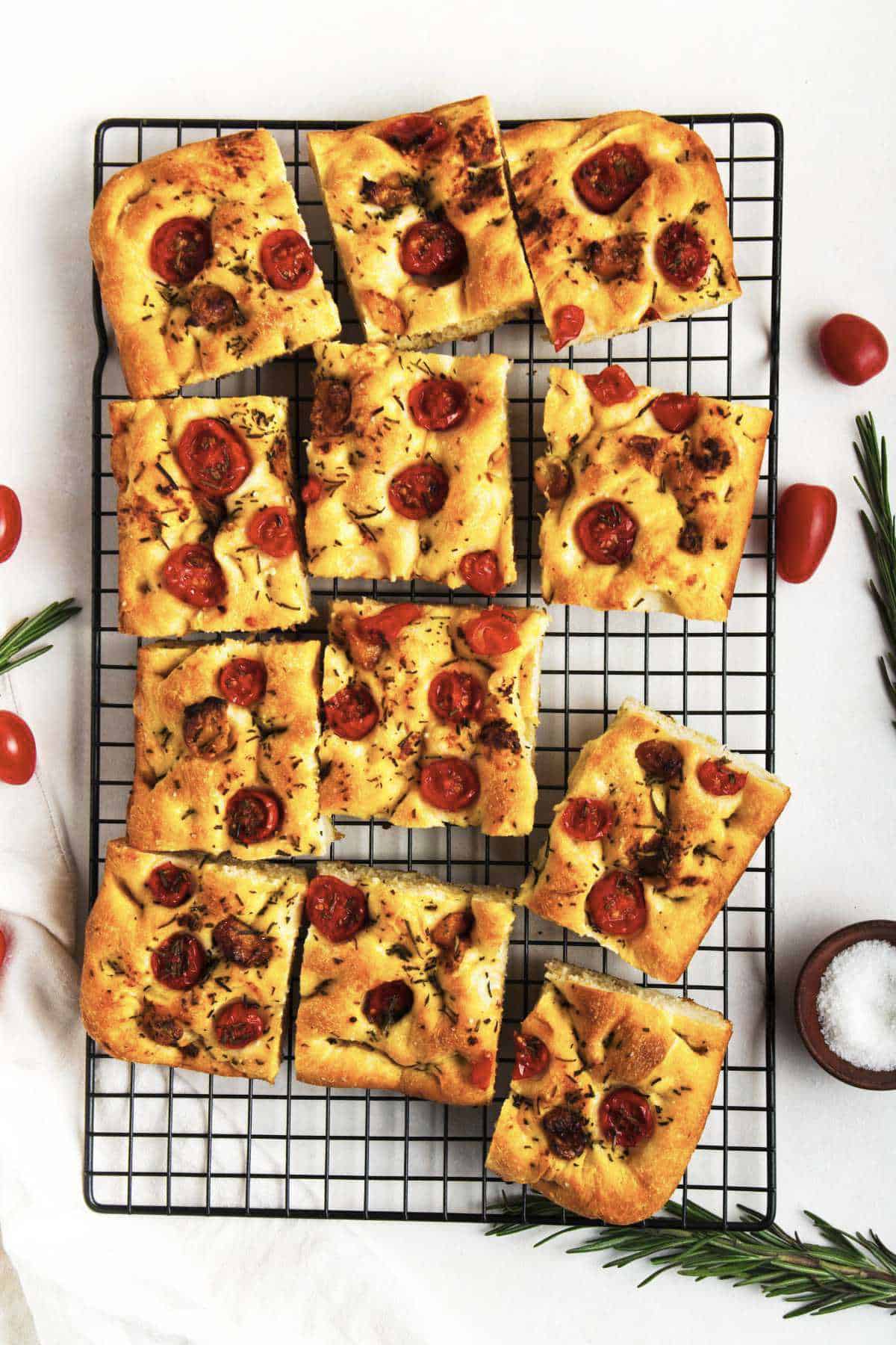 Sliced roasted garlic focaccia bread topped with cherry tomatoes and herbs on a black cooling rack, surrounded by rosemary sprigs, cherry tomatoes, and a small bowl of salt.