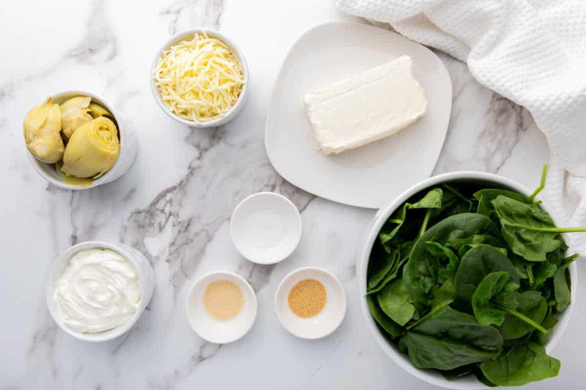 Ingredients for no mayo spinach artichoke dip arranged on a marble surface, including fresh spinach, artichoke hearts, shredded cheese, cream cheese, yogurt, and small bowls of spices, with a white cloth in the corner.
