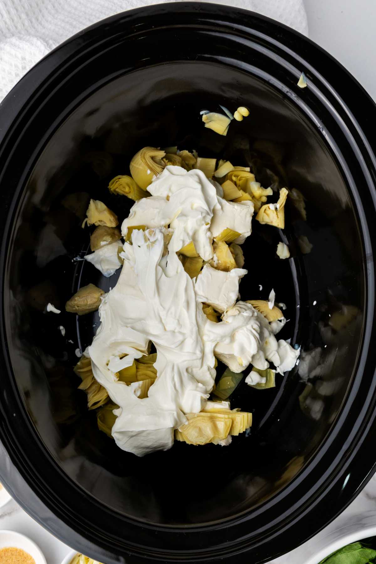 A close-up of a black slow cooker filled with artichoke hearts and cream cheese, ready to be mixed for a creamy no mayo spinach artichoke dip recipe.