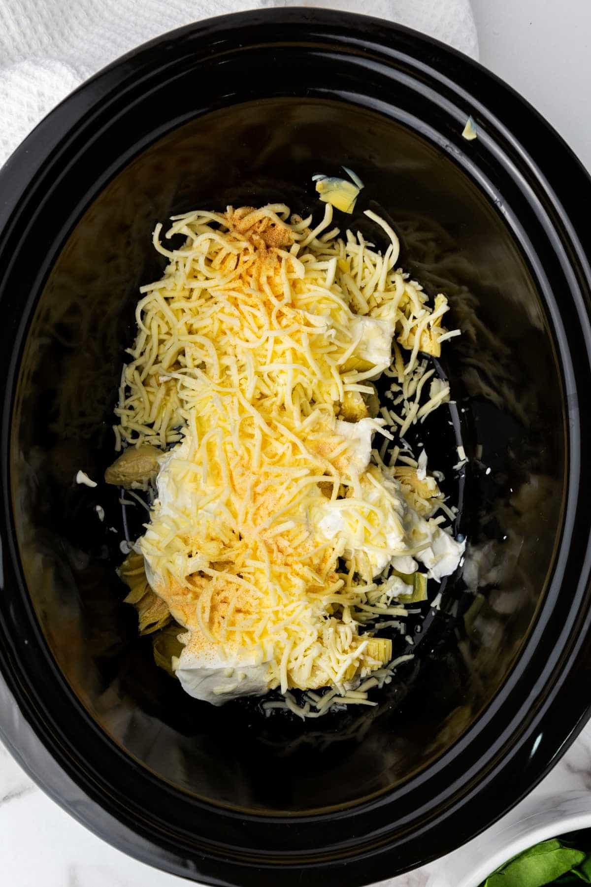 Shredded cheese, cream cheese, and seasonings are placed inside a black slow cooker for a no mayo spinach artichoke dip. A white cloth and part of a bowl are visible nearby, ready for serving.