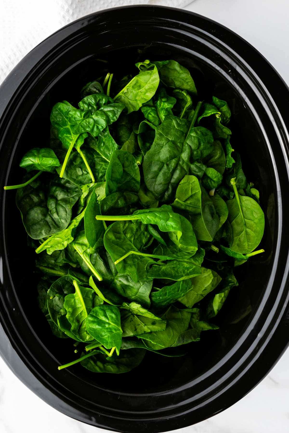 A black slow cooker filled with fresh, raw spinach leaves, viewed from above, ready to be transformed into a delicious no mayo spinach artichoke dip.
