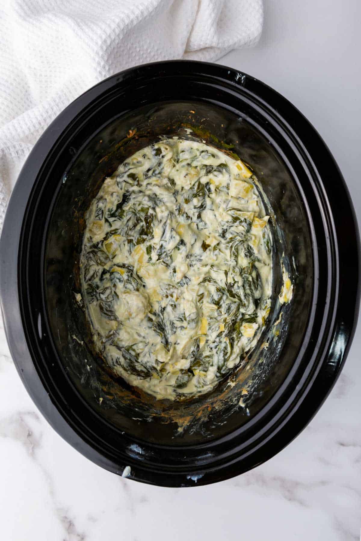 A slow cooker filled with creamy no mayo spinach artichoke dip sits on a white marble surface, with a white textured cloth partially visible in the background.