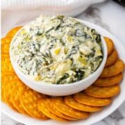 A white bowl filled with creamy no mayo spinach artichoke dip, surrounded by a ring of round, golden crackers on a white plate.