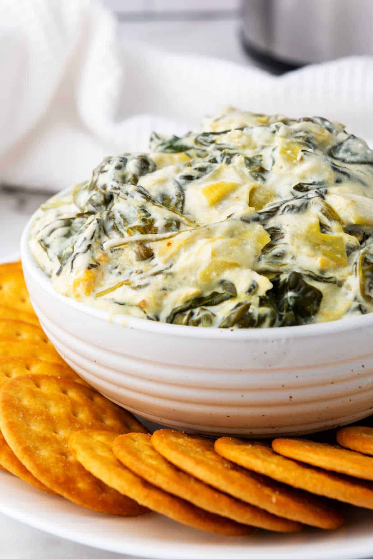 A white bowl filled with rich, cheesy no mayo spinach artichoke dip sits on a plate surrounded by round crackers. The dip appears creamy with visible spinach and artichoke pieces.