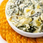 A white bowl filled with creamy no mayo spinach artichoke dip is surrounded by a ring of round golden crackers on a white plate.