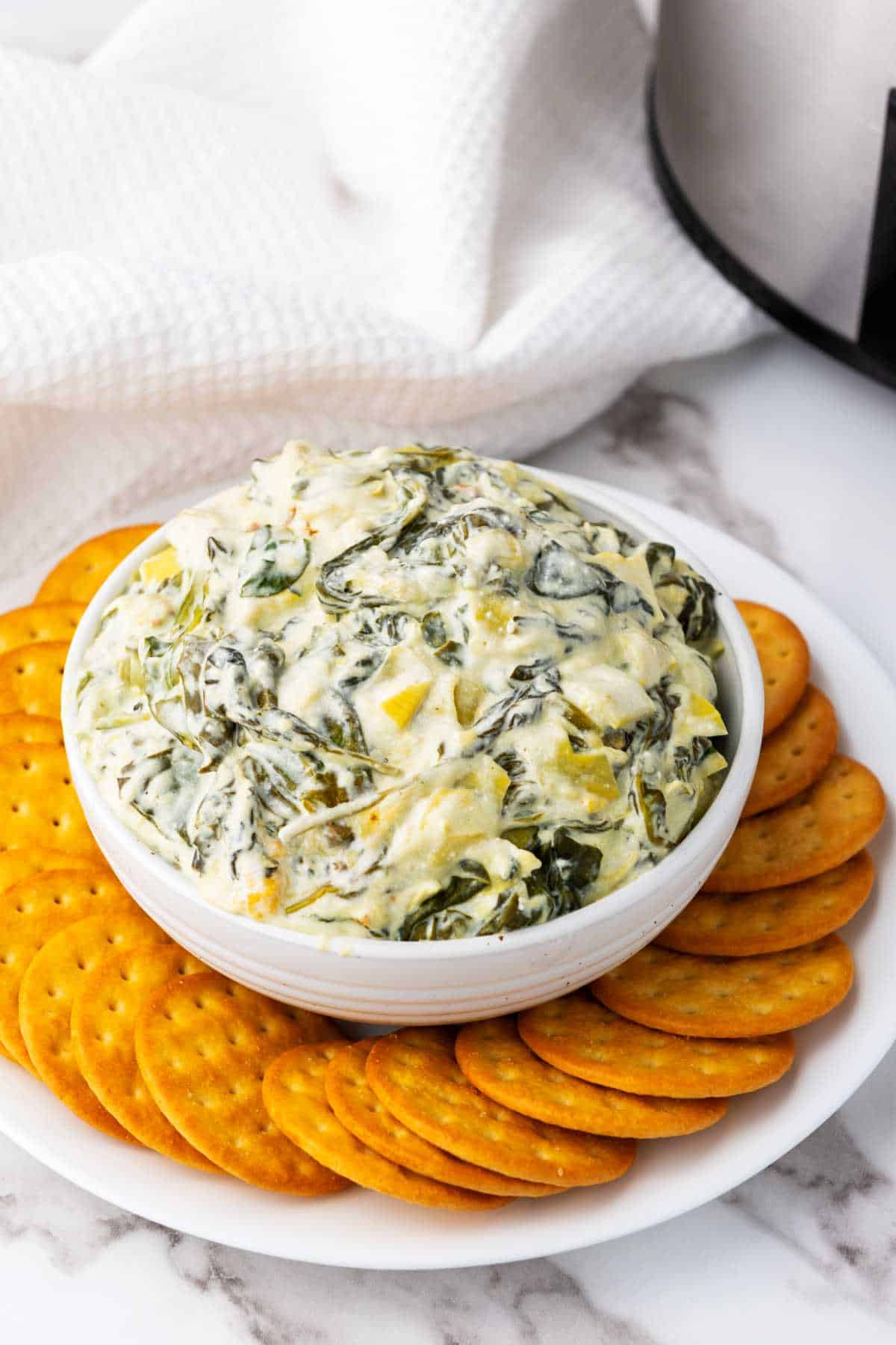 A white bowl filled with creamy no mayo spinach artichoke dip sits on a white plate, surrounded by round golden crackers. The plate is on a marble surface with a white textured cloth in the background.