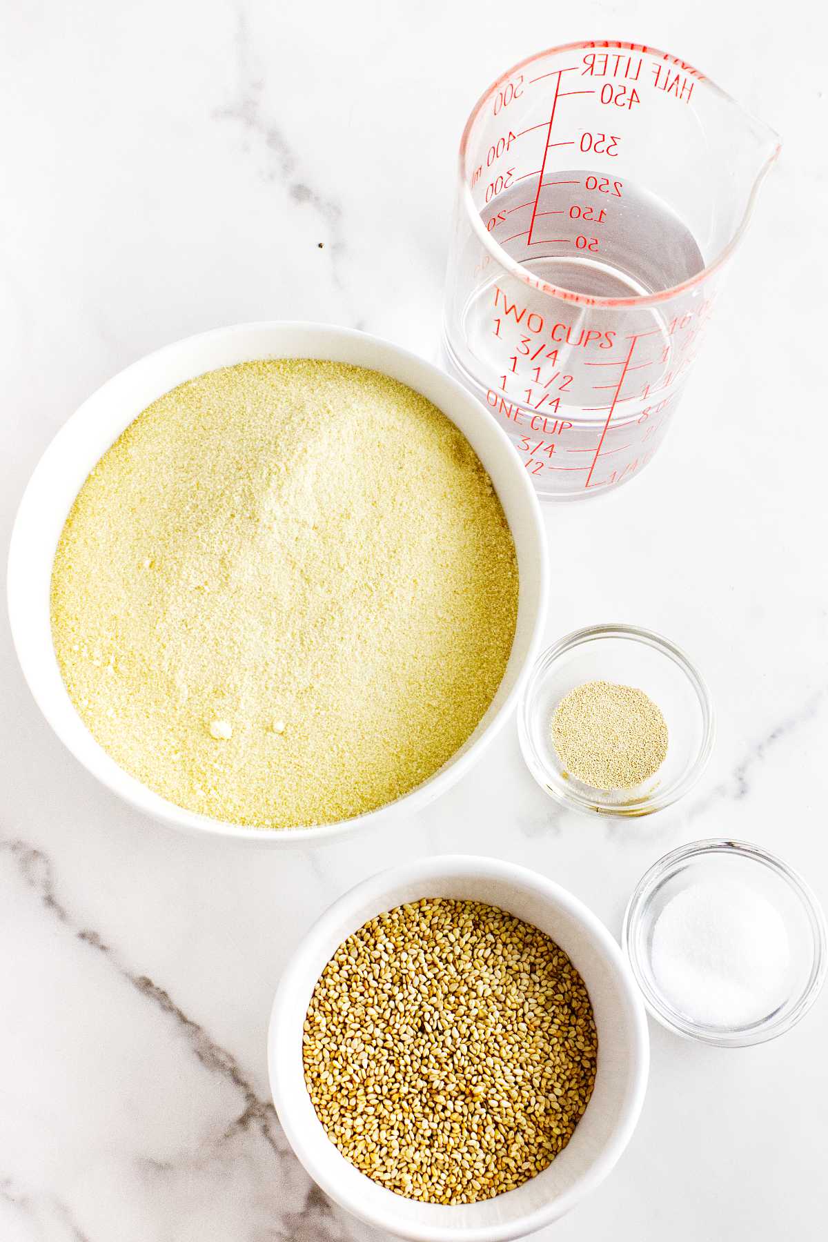 A measuring cup of water, a bowl of cornmeal, a small bowl of yeast, coarse corn grits, and salt are arranged on a white marble surface—perfect ingredients for making delicious semolina bread.