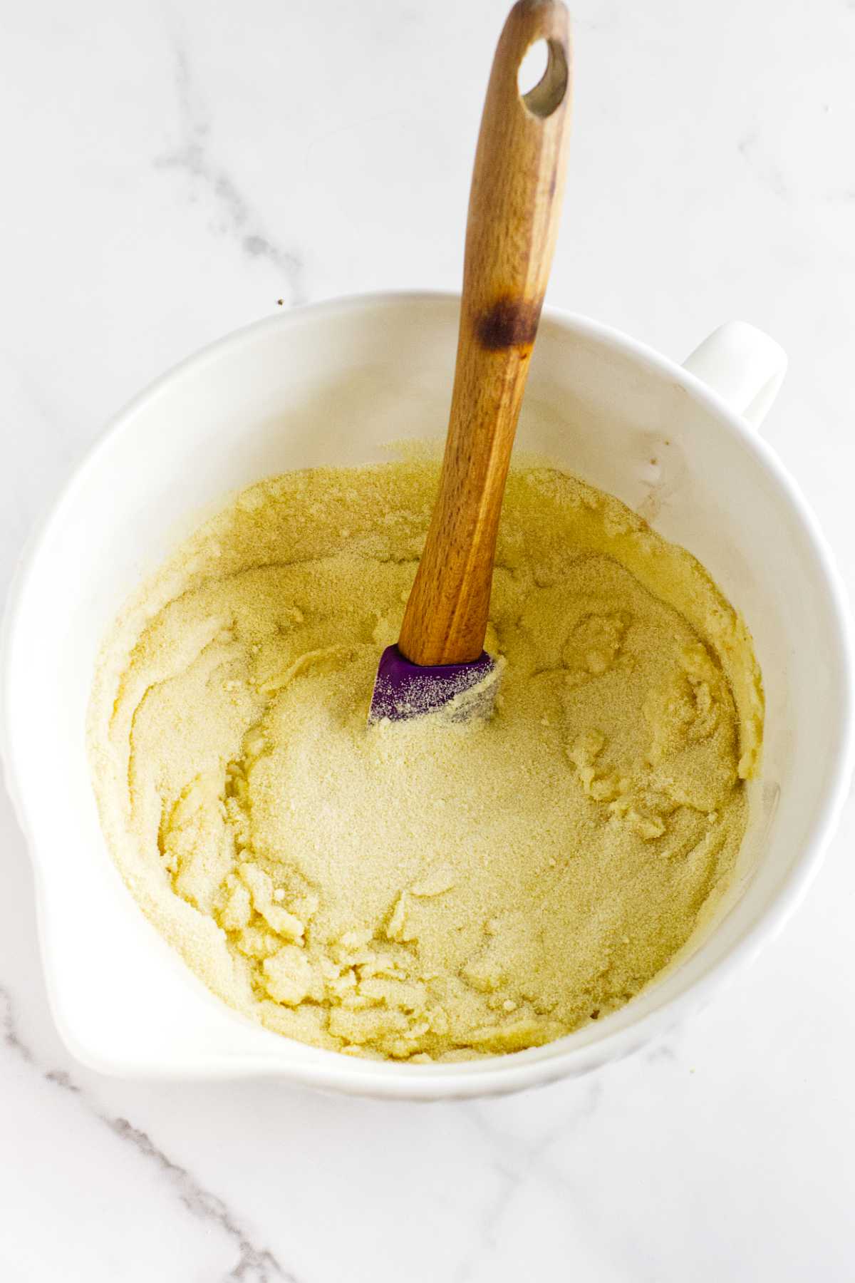 A white mixing bowl with yellow cornmeal mixture, perfect for making semolina bread, and a wooden spatula resting inside, placed on a white marble surface.