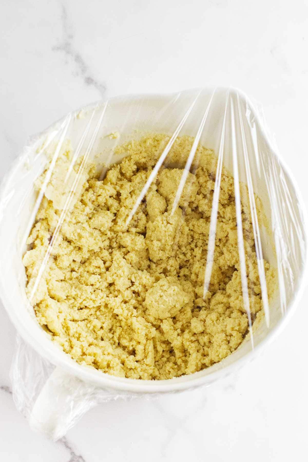 A white bowl filled with crumbly yellow semolina bread dough, covered with clear plastic wrap, sits on a white marble surface.