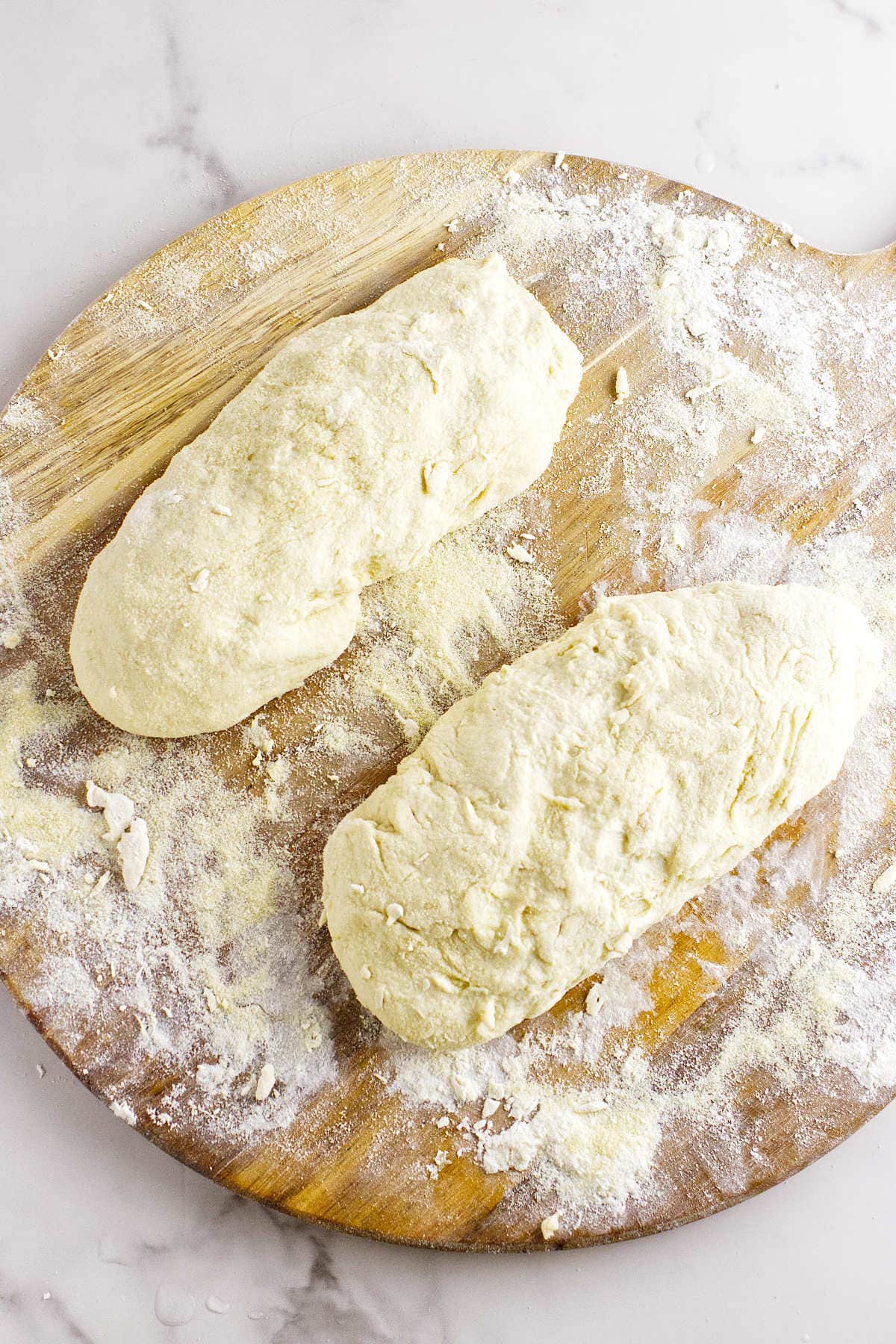 Two pieces of raw, floured semolina bread dough rest on a wooden board dusted with flour, ready for baking. The background is a white, marble-like surface.