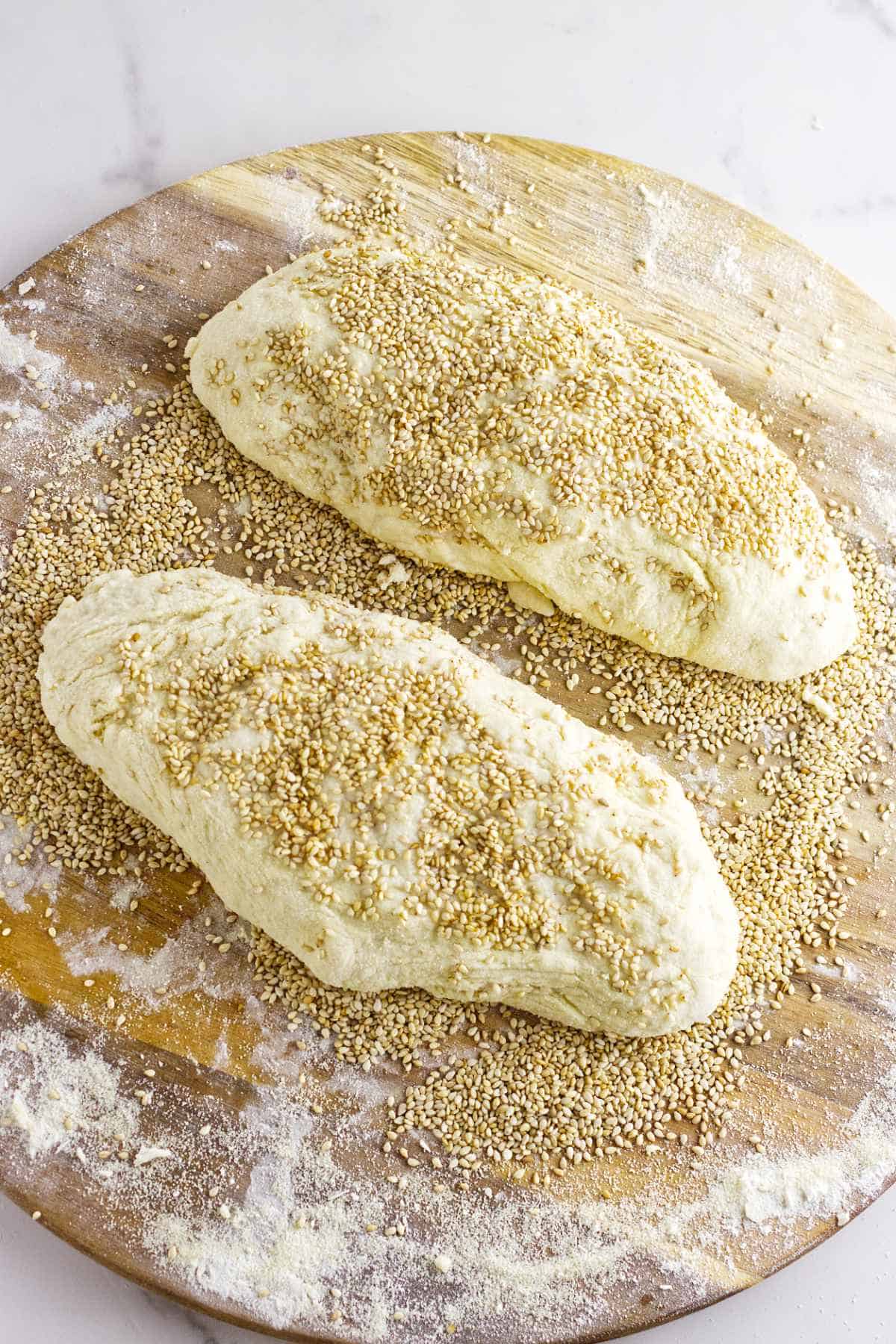 Two unbaked, oval-shaped loaves of semolina bread coated with sesame seeds rest on a floured wooden surface, ready for baking.