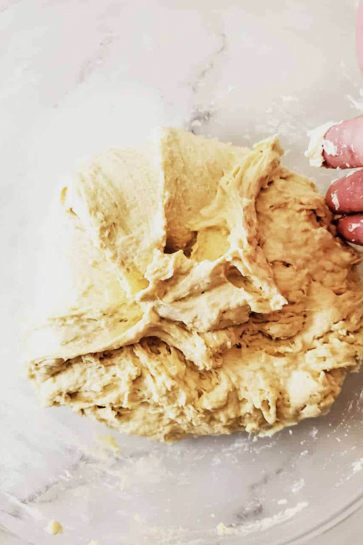 A close-up of a hand mixing a sticky, pale yellow semolina bread dough on a white, lightly floured surface. The soft, textured dough is ready for kneading or further preparation.