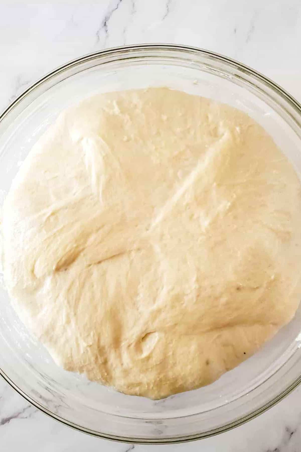 A glass bowl filled with risen, smooth pizza dough, perfect for making homemade semolina bread, sits on a white marble surface.