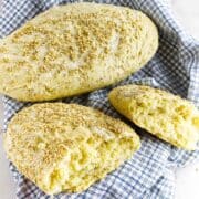 Two pieces of rustic semolina bread topped with sesame seeds, one whole and one broken open, rest on a blue and white checkered cloth.