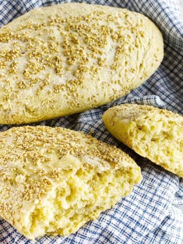 Two pieces of rustic semolina bread topped with sesame seeds, one whole and one broken open, rest on a blue and white checkered cloth.