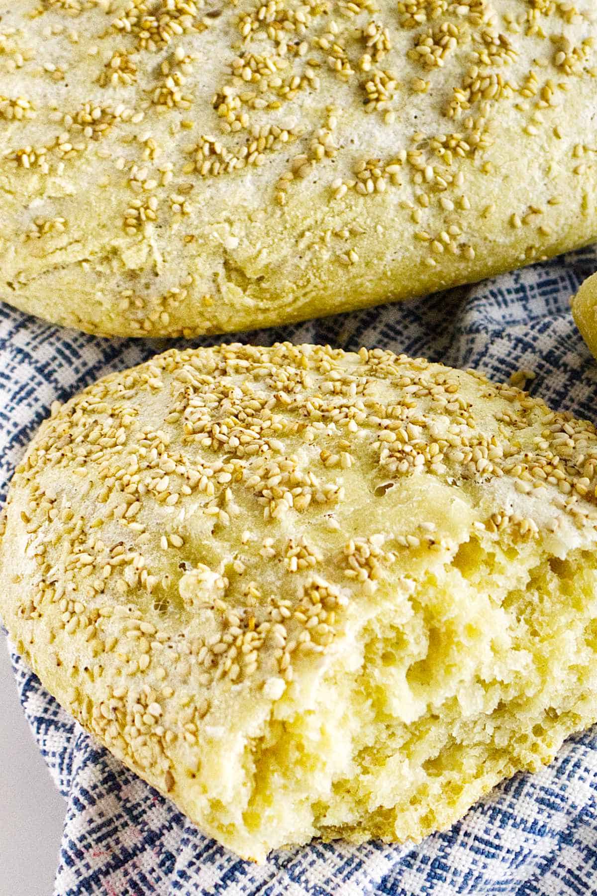 A close-up of freshly baked semolina bread topped with sesame seeds, with one loaf partially broken open to reveal its soft, fluffy interior. The bread rests on a blue and white patterned cloth.