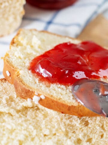 A spoon spreading vibrant strawberry rhubarb jam on a slice of fresh, white bread, with another slice and a loaf of bread in the background.