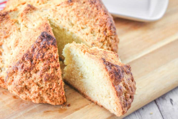 A round loaf of golden-brown Irish soda bread, made from a traditional Irish soda bread recipe, sits on a wooden cutting board with one wedge-shaped slice partially removed. The bread looks rustic with a crusty exterior and soft, crumbly interior.