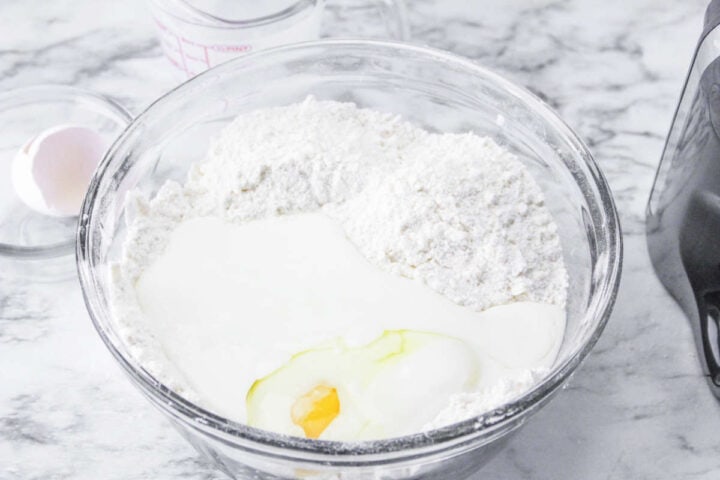 A clear glass bowl with flour, an egg, and milk on a marble countertop-perfect ingredients for a traditional Irish soda bread recipe-sits beside a whole egg, glass measuring cup, and black kitchen appliance.